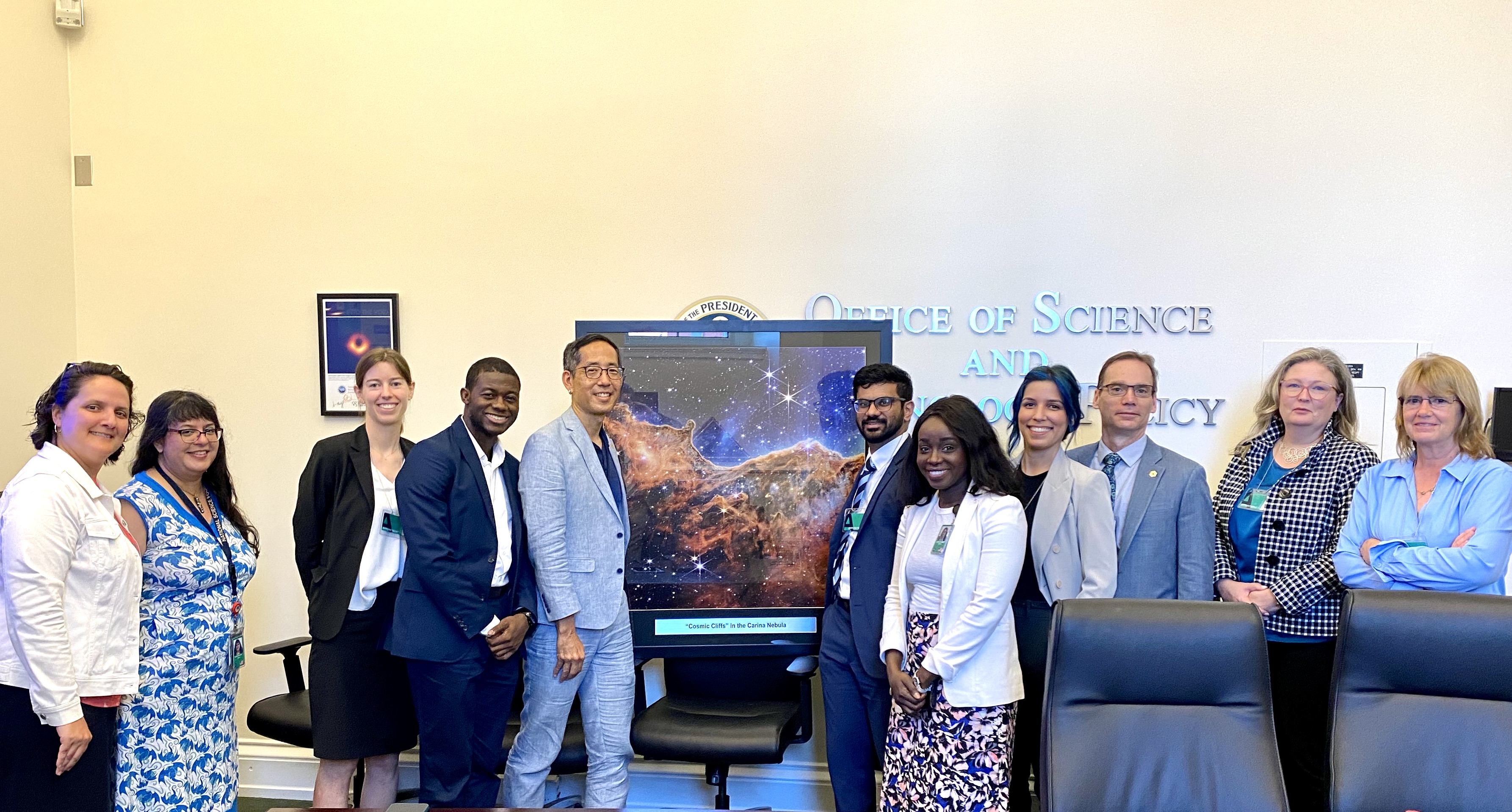 Photo of eleven people in suits in a conference room, posing with a framed photo of a nebula taken by JWST.